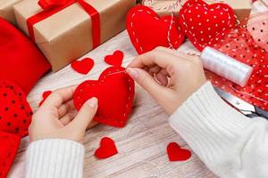 Valentine day theme. Workplace for preparing handmade decorations. Top view of female hands sew felt heart. Packed gifts, tools shabby wooden table photo