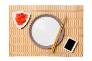 Empty round white plate with chopsticks for sushi and soy sauce, ginger on yellow bamboo mat background. Top view with copy space for you design photo