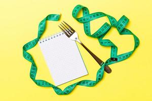 Top view of weight loss concept on yellow background. Mix of green centimeter tape, note pad and fork with space for your design photo