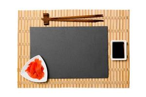 placa de pizarra negra rectangular vacía con palillos para sushi, jengibre y salsa de soja sobre fondo de estera de bambú amarillo. vista superior con espacio de copia para su diseño foto