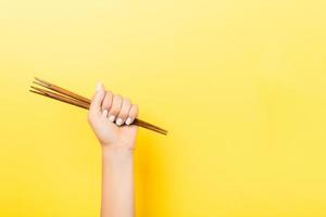 Cropped image of female hand holding chopsticks in fist on yellow background. Asian food concept with copy space photo
