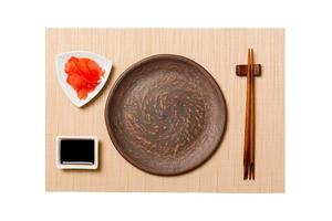 Empty round brown plate with chopsticks for sushi, ginger and soy sauce on brown sushi mat background. Top view with copy space for you design photo