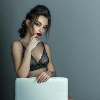 portrait of gorgeous woman with red lips and beautiful hairstyle sits on white leather chair in grey lace bra photo
