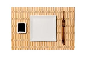 Empty white square plate with chopsticks for sushi and soy sauce on yellow bamboo mat background. Top view with copy space for you design photo