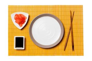 Empty round white plate with chopsticks for sushi, ginger and soy sauce on yellow bamboo mat background. Top view with copy space for you design photo