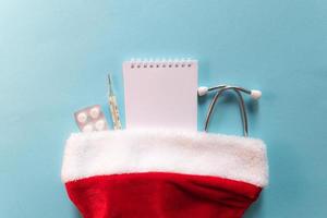 Medical Christmas and New year composition with stethoscope, pills, notepad and Santa hat on blue background. Health care and medicine concept for card, calendar, cover. photo