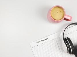 flat lay of habit tracker book, pink cup of coffee and headphones on white  background with copy space. photo