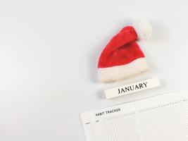 flat lay of habit tracker book, wooden calendar January, red Christmas Santa hat  on white  background with copy space. photo