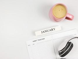 plano de libro de seguimiento de hábitos, calendario de madera enero, taza de café rosa y auriculares sobre fondo azul con espacio para copiar. foto