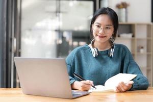 Young adult happy smiling Asian student wearing headphones talking on online chat meeting using laptop in university campus or at virtual office. College female student learning remotely. photo