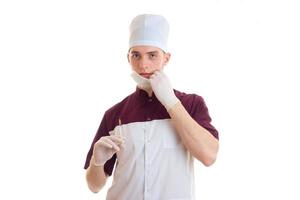 young honest doctor's hat and with a mask on his face looks straight and holding a syringe is isolated on a white background photo