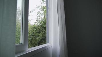 en un hotel o en una casa, el viento sopla y la naturaleza se puede ver a través de la ventana. el viento soplaba cortinas blancas que revoloteaban a través de la ventana abierta. video