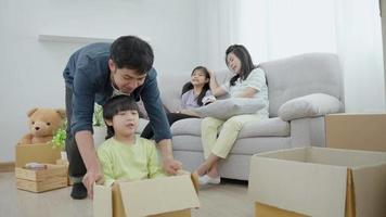 une jeune famille asiatique heureuse et souriante est en mouvement dans une nouvelle maison. le fils est assis dans une boîte en carton, son père déplace une boîte en carton. maman emmenant sa fille dans une nouvelle maison. video