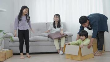 happy family is on the move to New room on new house. Son sit in a carton box and father moving box and have encouraged by daughter and wife. Moving home or house for family concept. video