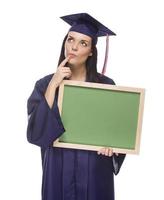 mujer graduada pensante en toga y birrete sosteniendo pizarra en blanco foto