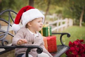 Young Child Wearing Santa Hat Sitting with Christmas Gifts Outside. photo