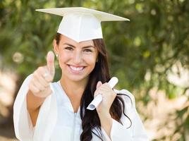 Mixed Race Thumbs Up Girl Celebrating Graduation Outside In Cap and Gown with Diploma in Hand photo