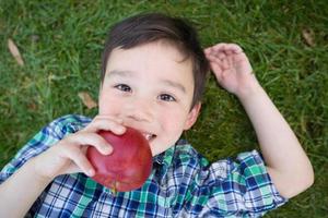 Mixed Race Chinese and Caucasian Young Boy With Apple Relaxing On His Back Outside On The Grass. photo