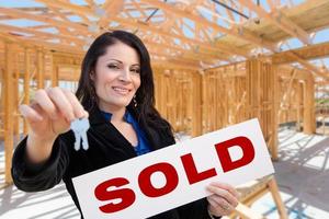 Hispanic Woman With Keys and Sold Sign On Site Inside New Home Construction Framing. photo