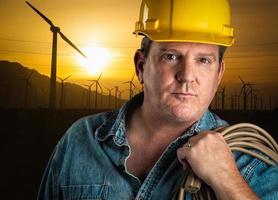 Serious Contractor in Hard Hat Holding Extension Cord Outdoors Near Wind Turbines. photo