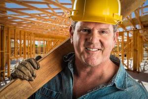 contratista sonriente con casco que sostiene un tablón de madera en el sitio de construcción.