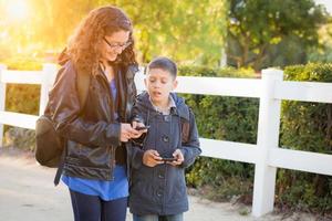 hermano y hermana hispanos con mochilas caminando enviando mensajes de texto en teléfonos celulares foto