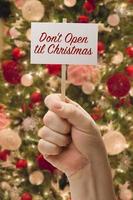 mano sosteniendo no abra hasta la tarjeta de navidad frente al árbol de navidad decorado. foto