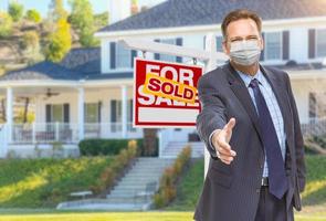 Male Real Estate Agent Reaching for Hand Shake Wearing Medical Face Mask with Sold For Sale Sign Behind photo