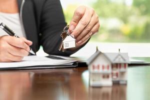 Woman signing real estate contract papers holding house keys and home keychain with small model home in front. photo