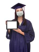 Graduating Female Wearing Medical Face Mask and Cap and Gown  Holding Blank Computer Tablet Isolated on a White Background photo