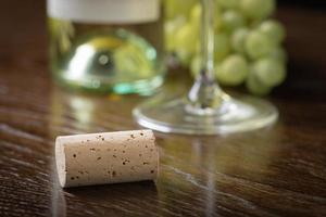 corcho de vino en blanco descansando sobre una mesa de madera cerca de uvas, botella y vidrio. foto
