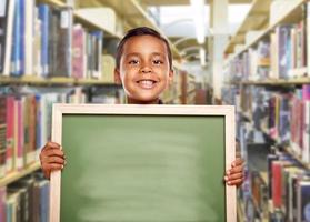 niño hispano sonriente sosteniendo una pizarra vacía en la biblioteca foto