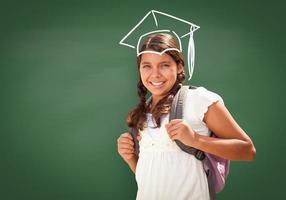 Young Hispanic Student Girl Wearing Backpack Front Of Blackboard with Fireman Helmet Drawn In Chalk Over Head photo