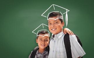 Young Hispanic Student Boys Wearing Backpacks In Front Of Blackboard with Graduation Caps Drawn In Chalk Over Head photo