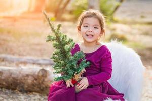 Linda niña de raza mixta sosteniendo un pequeño árbol de navidad al aire libre foto