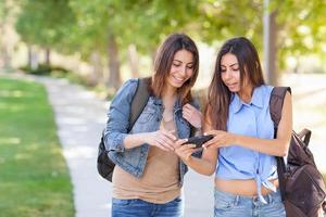 dos hermosas jóvenes hermanas gemelas étnicas con mochilas usando un teléfono inteligente afuera. foto