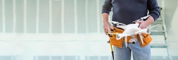 Construction Worker and Drone Pilot With Toolbelt Holding Drone In Front of Drywall and Ladder Banner photo