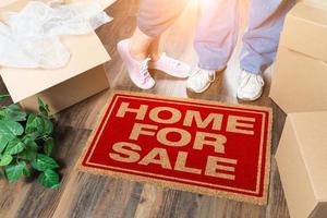 Man and Woman Standing Near Home For Sale Welcome Mat, Moving Boxes and Plant photo