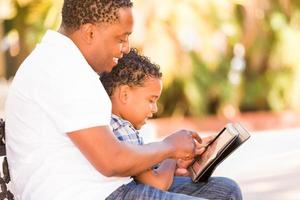 African American Father and Mixed Race Son Using Computer Tablet on Bench in Park photo