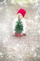 chica con mitones rojos y gorra cerca de un pequeño árbol de navidad con efecto de nieve foto