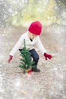 chica con mitones rojos y gorra cerca de un pequeño árbol de navidad con efecto de nieve
