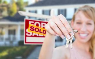 Excited Woman Holding House Keys and Sold For Sale Real Estate Sign in Front of Nice New Home. photo