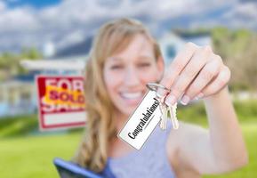 Woman Holding New House Keys with Congratulations Card In Front of Sold Real Estate Sign and Home. photo