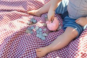 Baby Boy sentado en una manta para picnic poniendo monedas en la alcancía foto