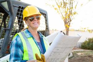 Smiling Female Worker Holding Technical Blueprints Near Small Bulldozer At Construction Site photo