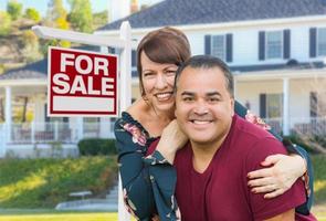 Pareja adulta joven de raza mixta en frente de la casa y en venta signo de bienes raíces foto