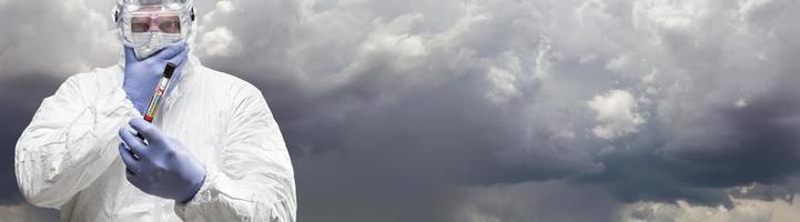 Male Doctor or Nurse In Hazmat Gear Holding Positive Coronavirus Test Tube Cloudy Sky Banner photo