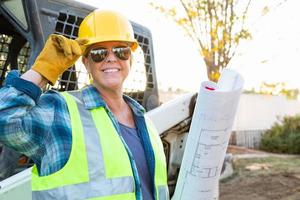 trabajadora sonriente sosteniendo planos técnicos cerca de una pequeña excavadora en el sitio de construcción foto