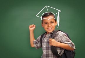 joven estudiante hispano con mochila frente a la pizarra con gorra de graduación dibujada con tiza sobre la cabeza foto
