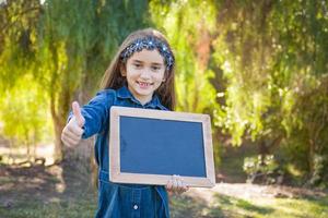 Cute Young Mixed Race Girl With Thumbs Up Holding Blank Blackboard Outdoors photo